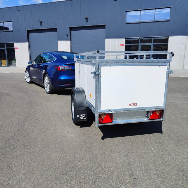 Witte bagage aanhangwagen met dakdragers op het kofferdeksel om op reis te gaan met de auto. Bovenop het deksel kunnen dakdragers gemonteerd worden voor een extra fietsenrek of dakkoffer. De bagagewagen hangt aan een elektrische auto merk Tesla klaar om te vertrekken op reis