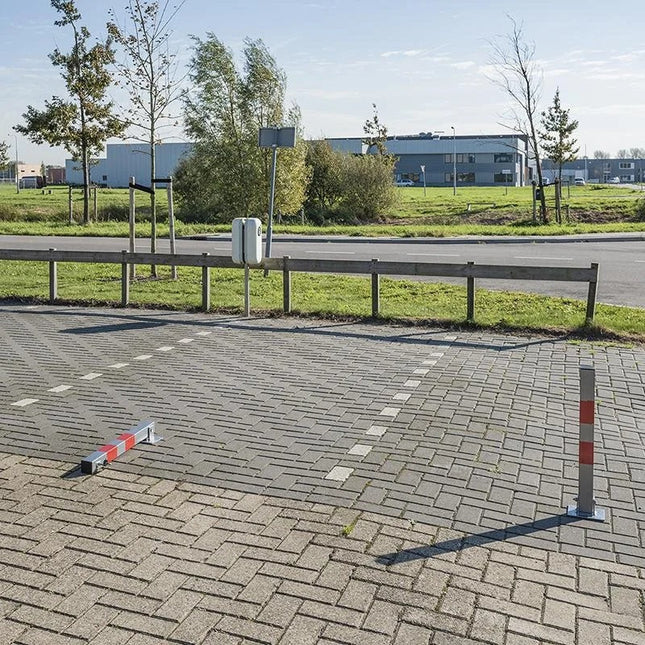 Grijze anti-parkeerpaal met rode reflecterende band. Bovenaan de parkeerpaal is een slot voor de sleutel. Onderaan is een bodemplaat om te verankeren aan de parking of grond. De paal kan onderaan scharnieren of kantelen om weg te rijden met de auto of aanhangwagen.